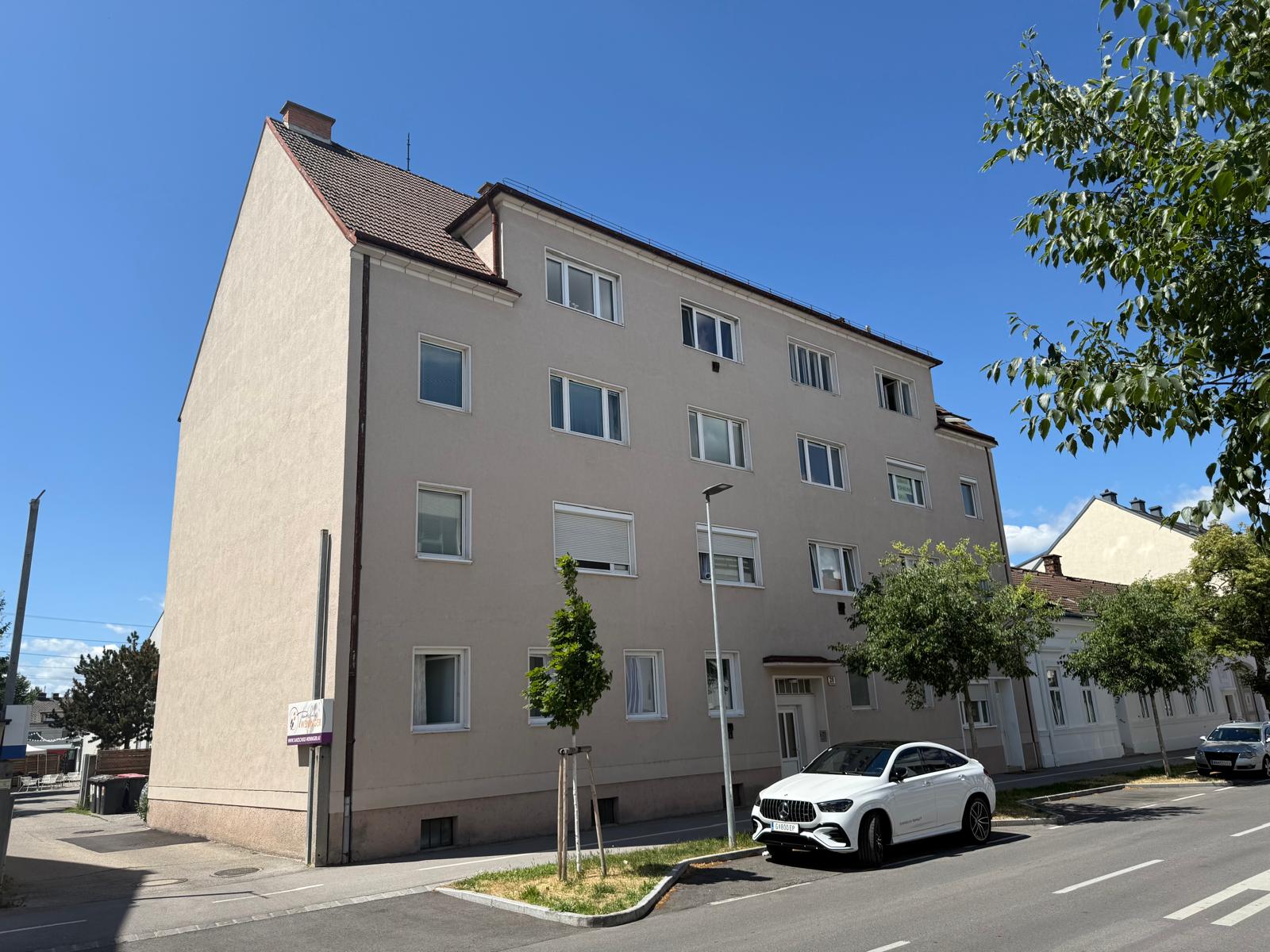 Wohnung in Wr. Neustadt, Haus mit beiger Fassade, Fenster weiß, Straße im Vordergrund, weißer Mercedes parkt davor, blauer Himmel