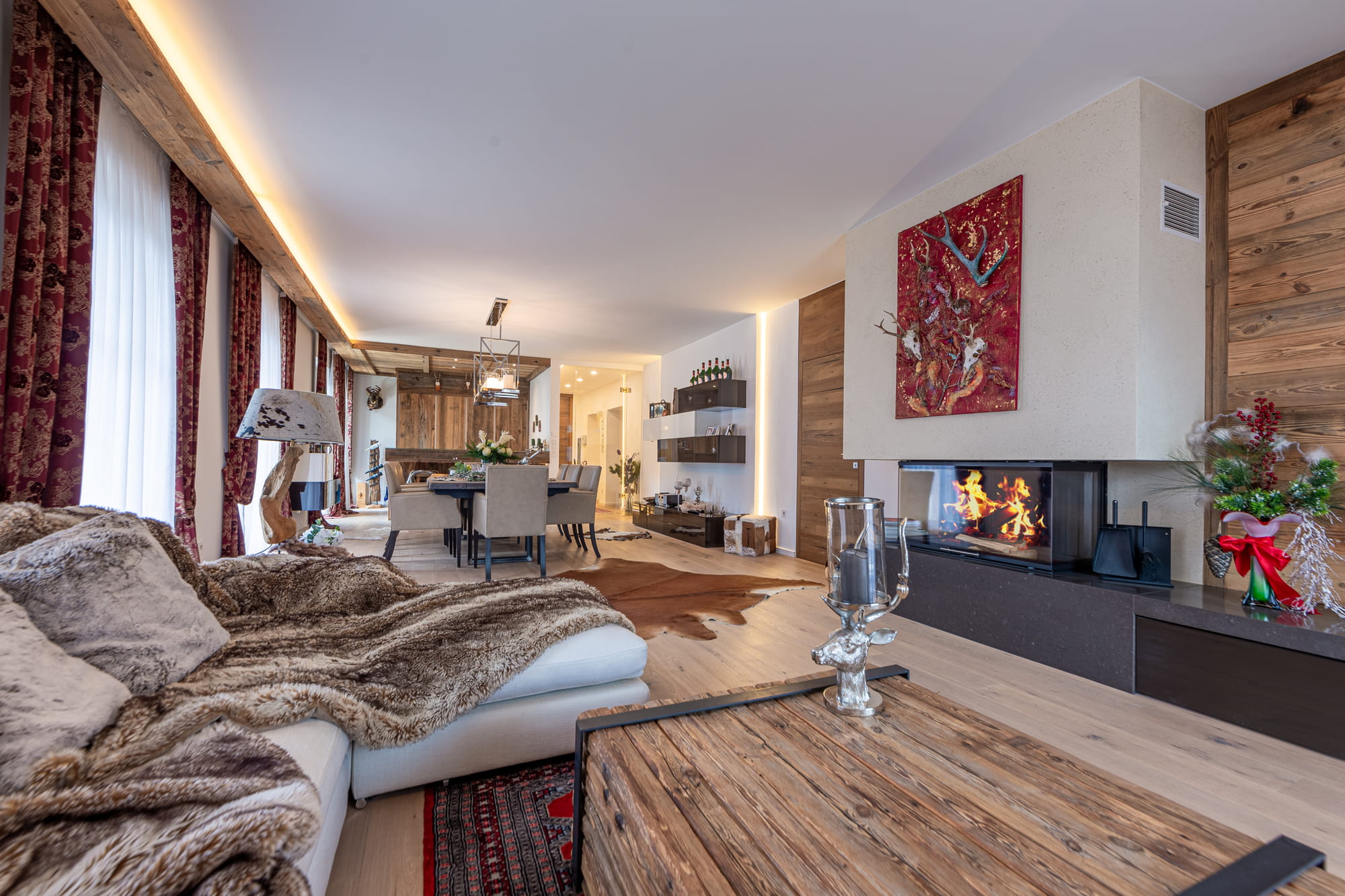 sehr schöne, luxuriöse Wohnung mit alpinem Charme in Kitzbühel, großzügiger Wohn- und Essraum, mit Holzboden, Sofa mit Couchtisch und offener Kamin im Vordergrund, dahinter schöner Essbereich