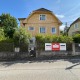 Einfamilienhaus in Wien-Hietzing, Straße im Vordergrund, Vorgarten, Haus mit gelber Fassade, weiße Fenster, Verkaufsbanner der Firma Helion am Gartenzaun