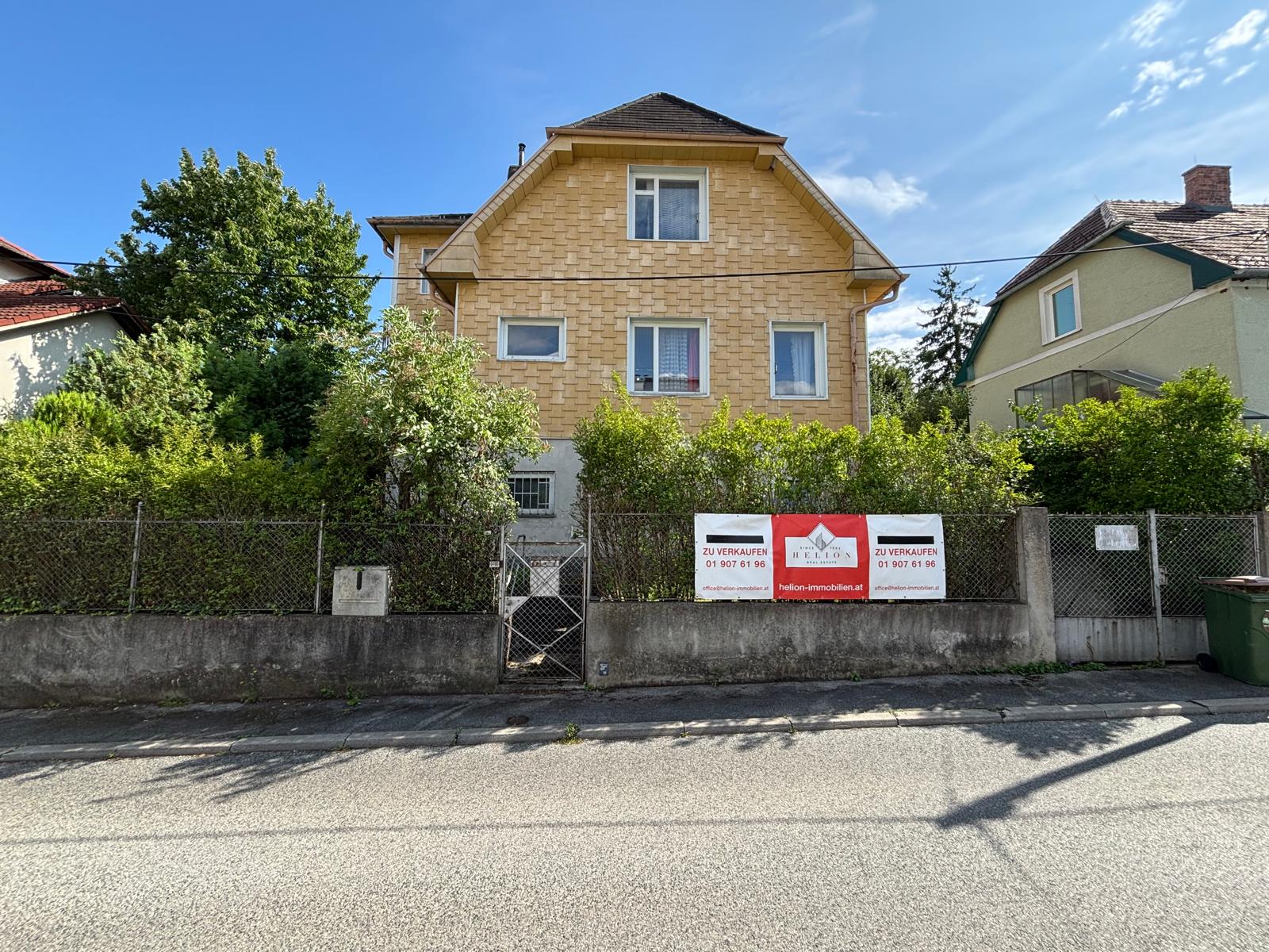 Einfamilienhaus in Wien-Hietzing, Straße im Vordergrund, Vorgarten, Haus mit gelber Fassade, weiße Fenster, Verkaufsbanner der Firma Helion am Gartenzaun