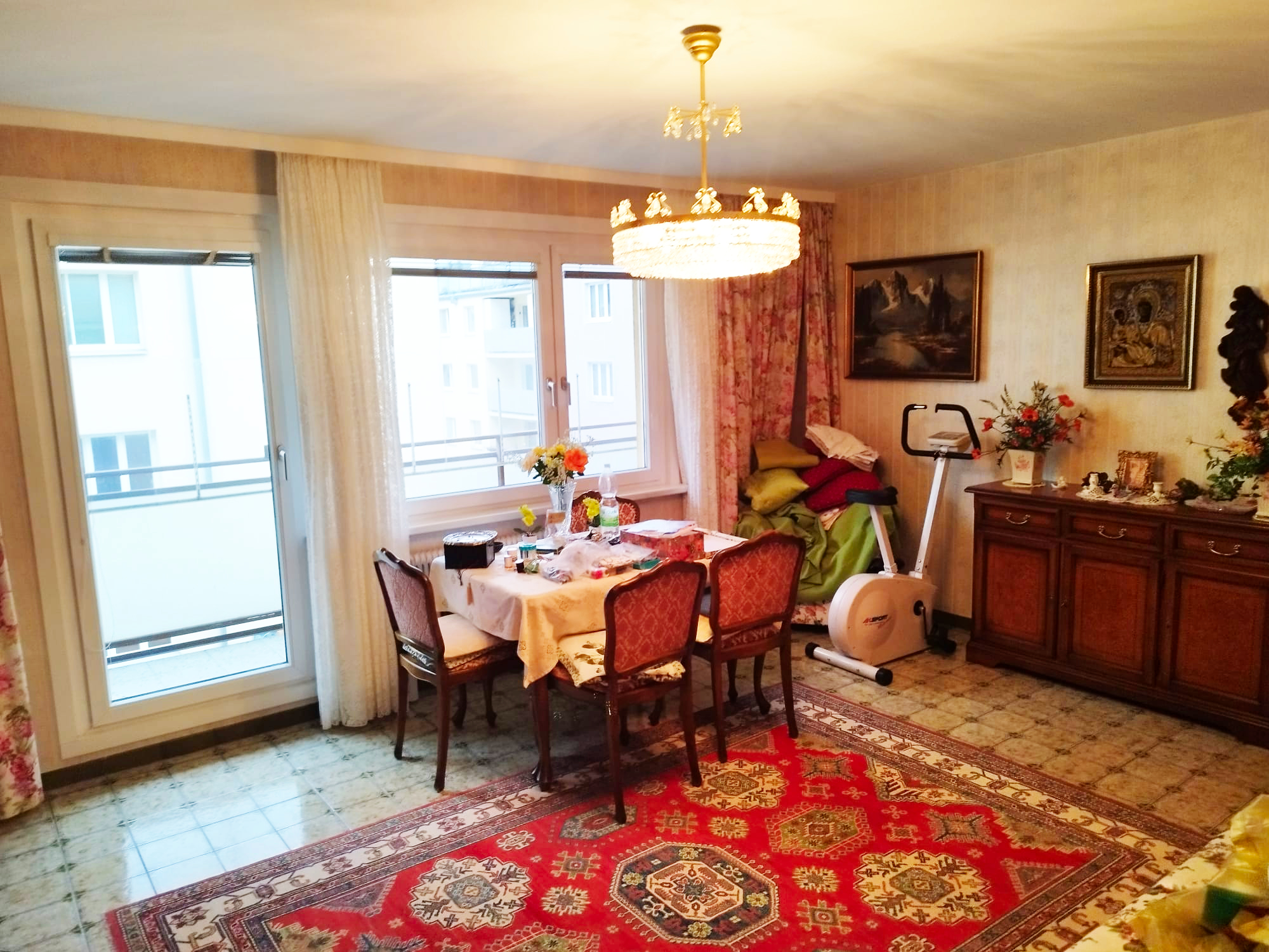 Wohnzimmer mit Ausgang auf die Terrasse und Fenster, rot gemusterter Teppich, Esstisch mit 4 Sessel, Anrichte aus dunklem Holz, Kronleuchter an der Decke, 2 "antike" Bilder rechts an der Wand, 5. Bezirk in Wien