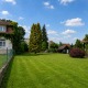 wunderschöner Baugrund in 1130 Wien, große gepflegte Rasenfläche, links Nachbarhaus, 3 hohe Bäume im Hintergrund, Strauch rechts vorne, dahinter ein kleines Holzhaus, blauer Himmel mit vereinzelt Wolken