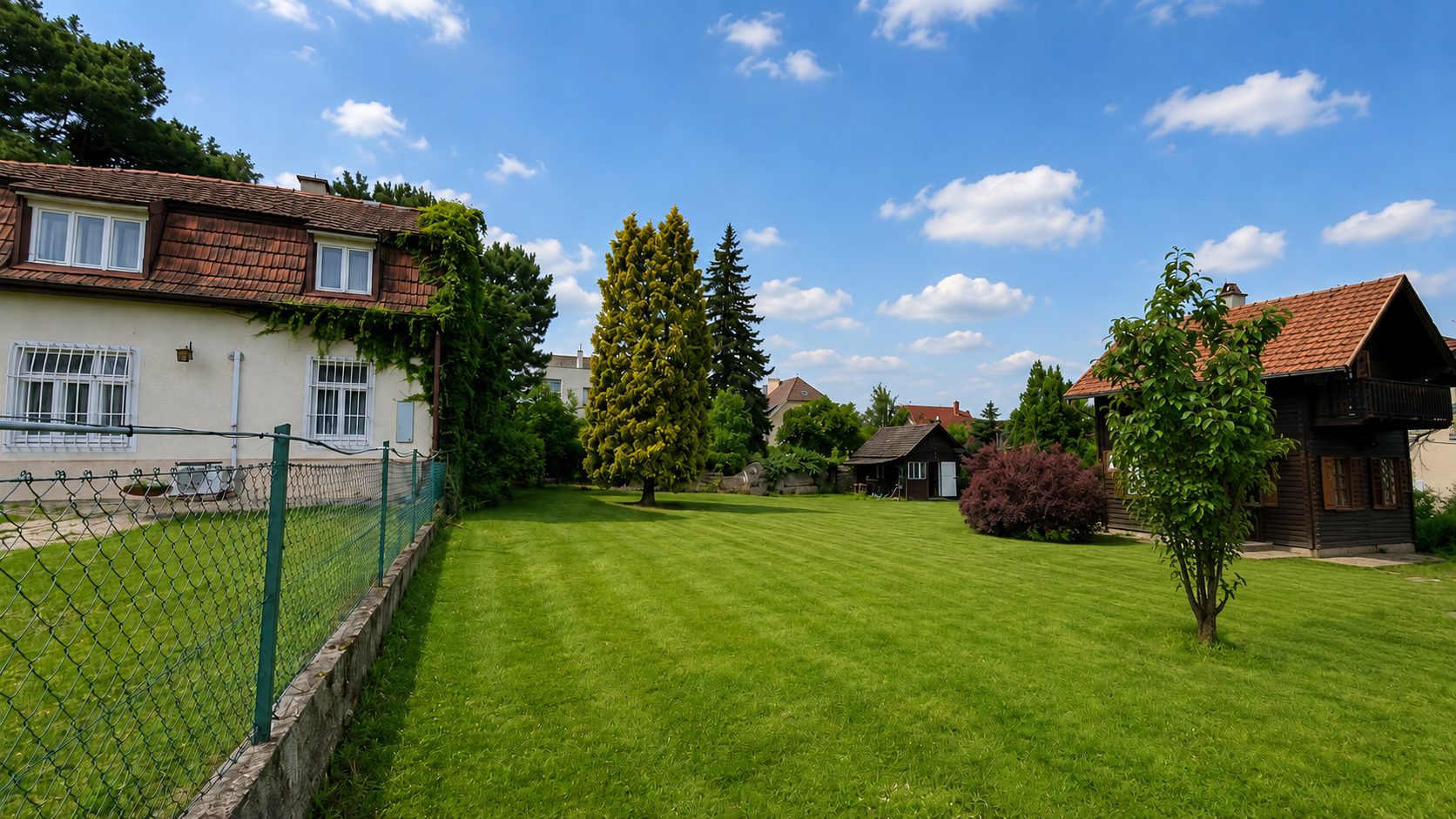 wunderschöner Baugrund in 1130 Wien, große gepflegte Rasenfläche, links Nachbarhaus, 3 hohe Bäume im Hintergrund, Strauch rechts vorne, dahinter ein kleines Holzhaus, blauer Himmel mit vereinzelt Wolken