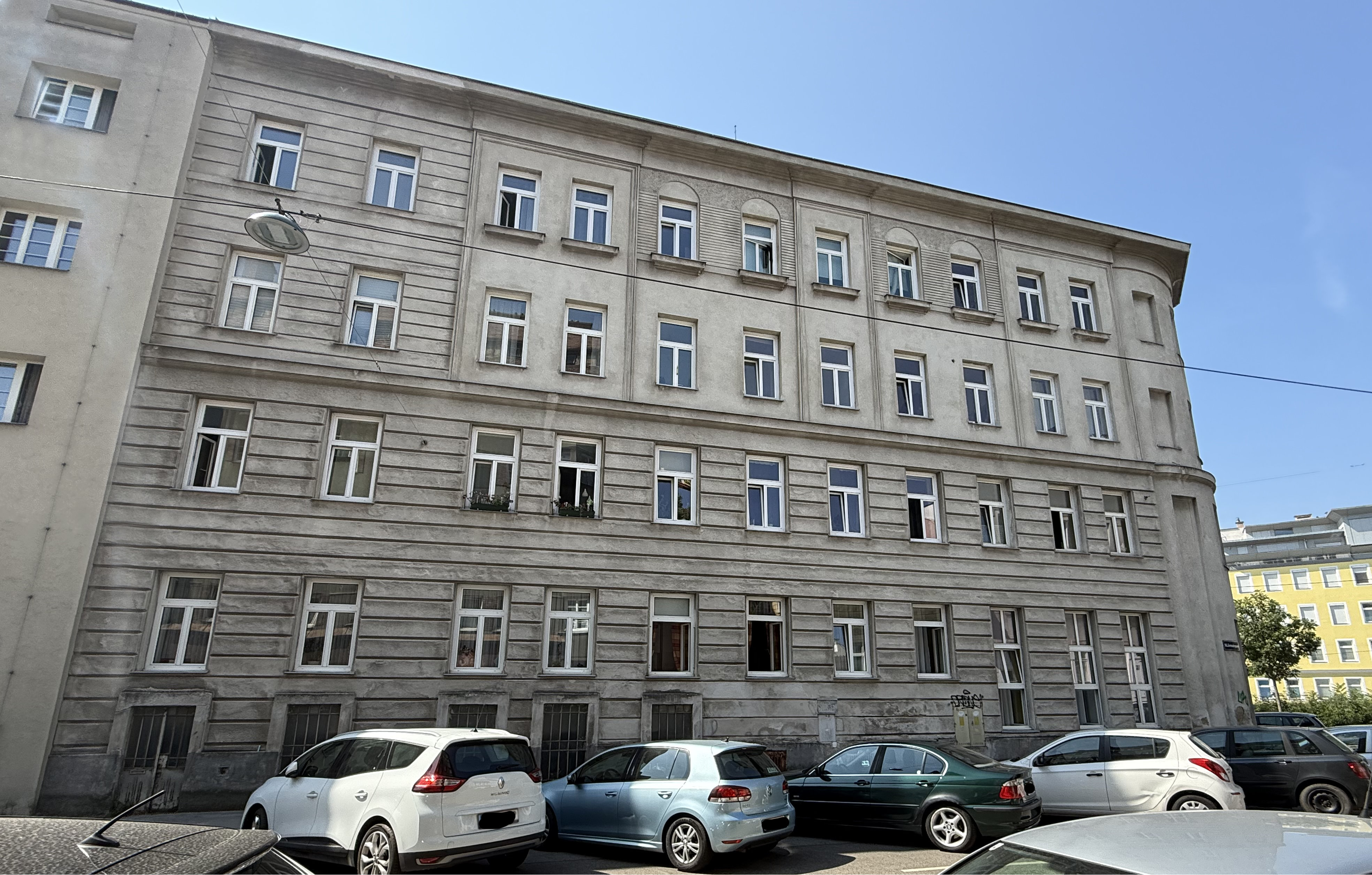 Blick auf ein Eckzinshaus im 14. Bezirk von Wien, blauer Himmel, graue Fassade, weiße Fenster