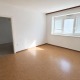 heller unmöbliertes Zimmer, mit weißen Wänden und Fensterfront mit Jalousien, rot/brauner Fußboden, Durchgang in einen anderen Raum