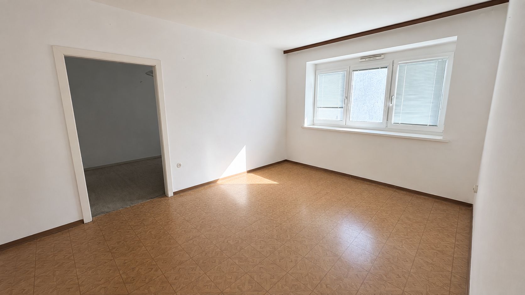heller unmöbliertes Zimmer, mit weißen Wänden und Fensterfront mit Jalousien, rot/brauner Fußboden, Durchgang in einen anderen Raum